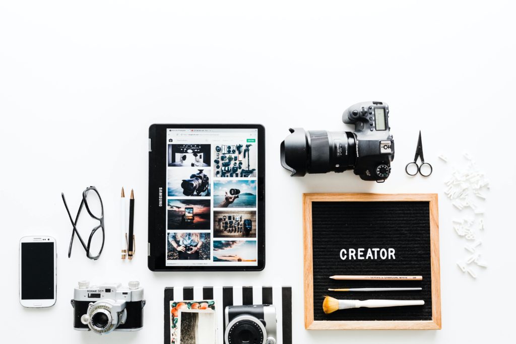 Biurko fotografa-artysty: obok siebie ułożone aparaty, notes, telefon, okulary i tablet z otwartą stroną internetową ze zdjęciami.