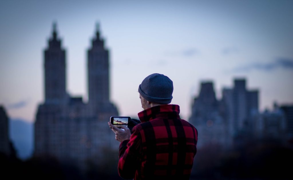 Na fotografii znajduje się mężczyzna, który zastanawia się, jak robić lepsze zdjęcia telefonem. Jego sylwetka skierowana jest w kierunku panoramy miasta. 