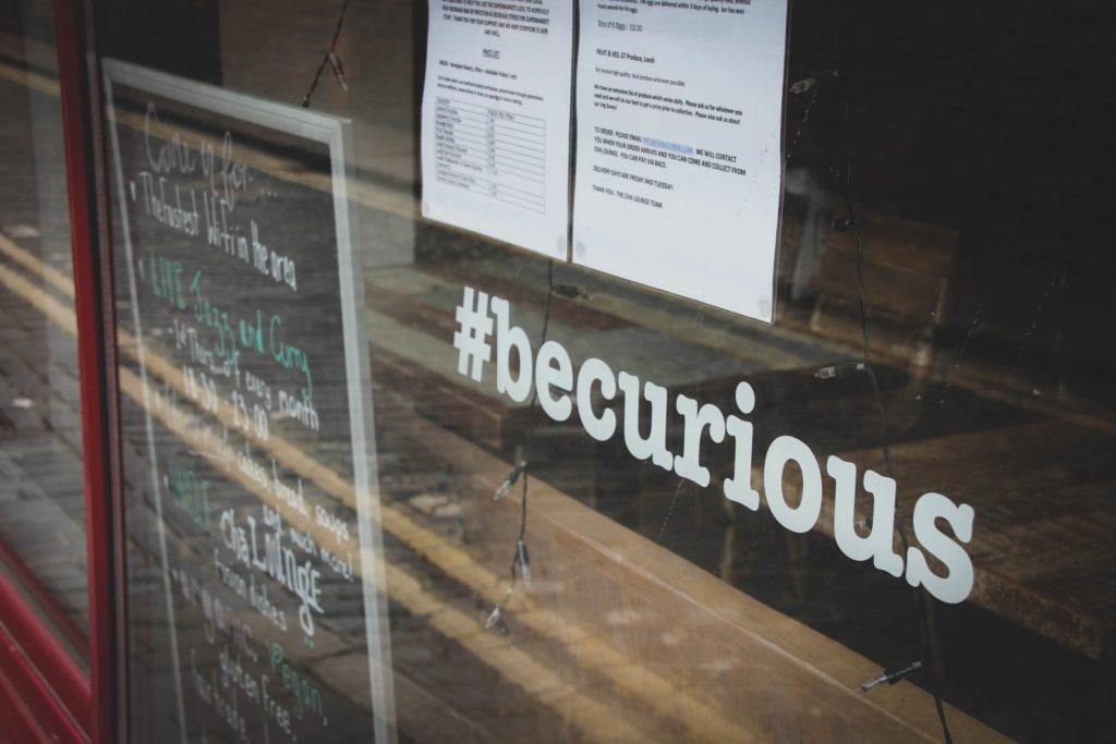 Obraz przedstawia witrynę sklepową, z hashtagiem zawierającym określenie: becurious. Obok widać tablicę z menu oraz inne ogłoszenia wywieszone na witrynie.